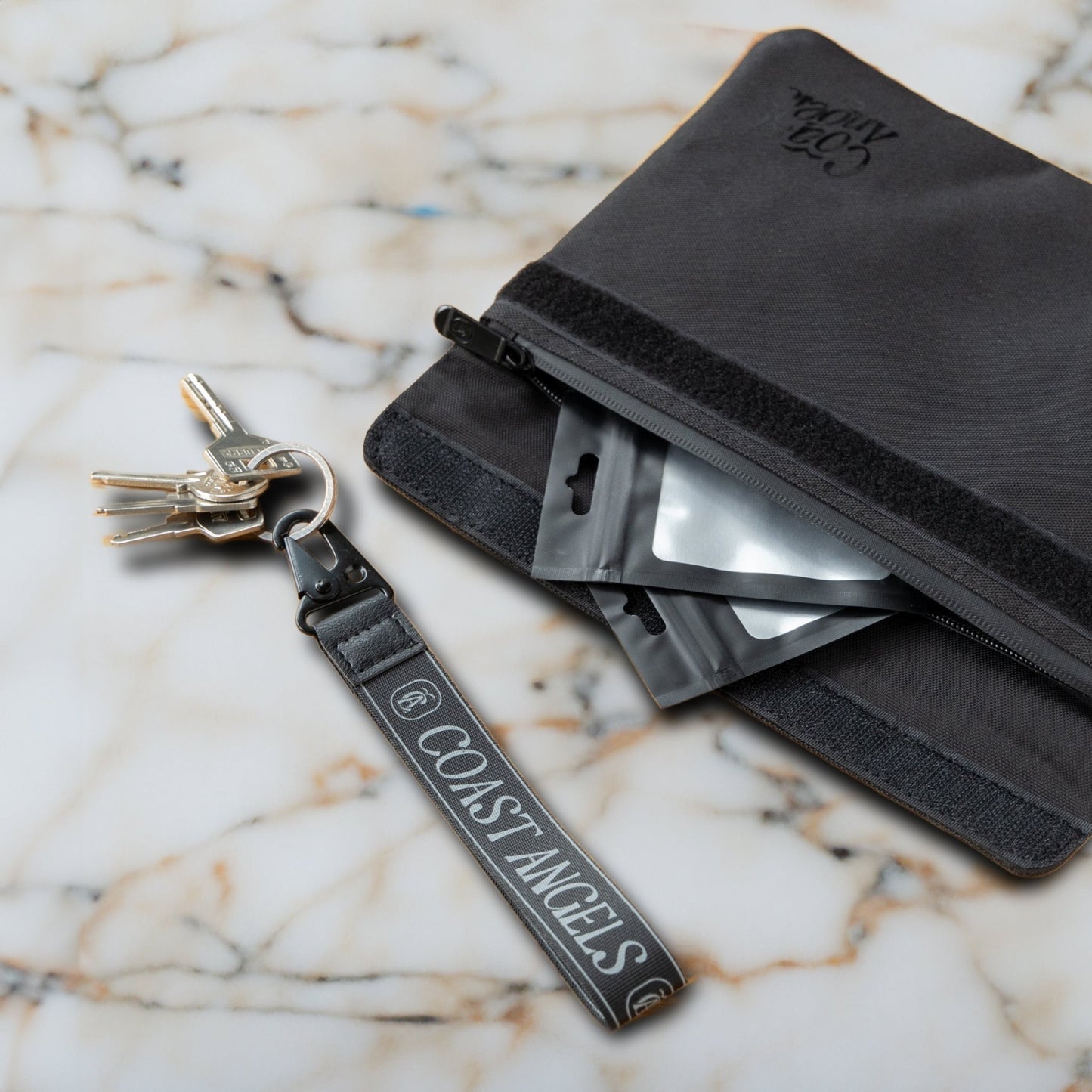 Black Coast Angels Soho smell-proof pouch shown partially unzipped on a marble surface, with resealable bags visible inside and a branded wrist strap holding a set of keys — showcasing portable, discreet functionality.